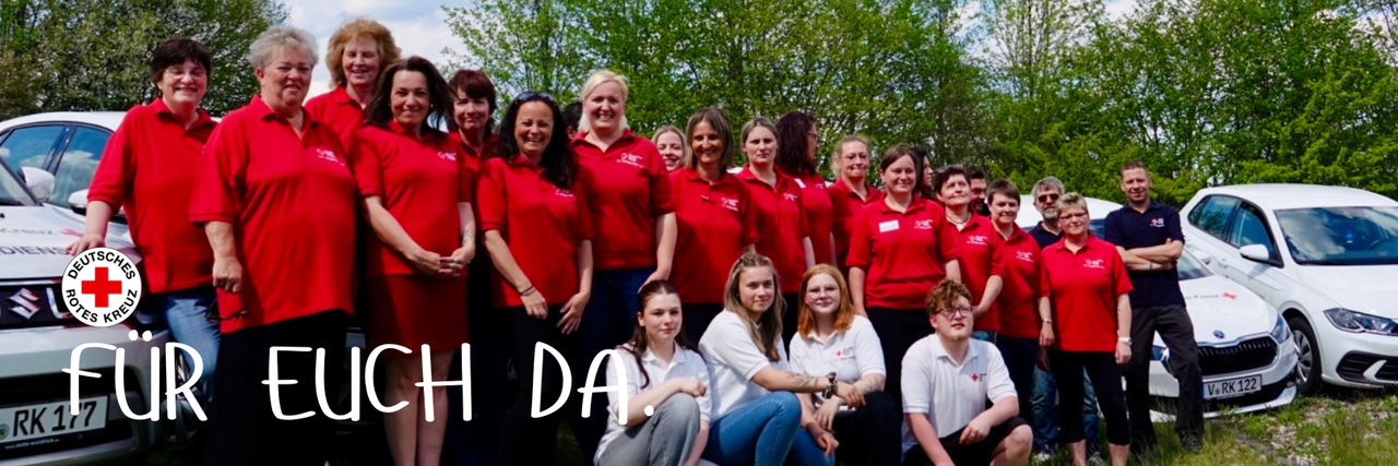 Die Mitarbeitenden unserer DRK Sozialstation stehen in roten Poloshirts vor ihren Autos, davor, hockend, in weißen Poloshirts unsere Azubis.